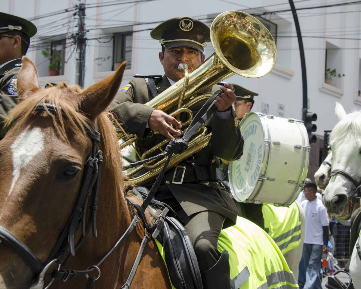A military band member plays a large horn while riding his horse
