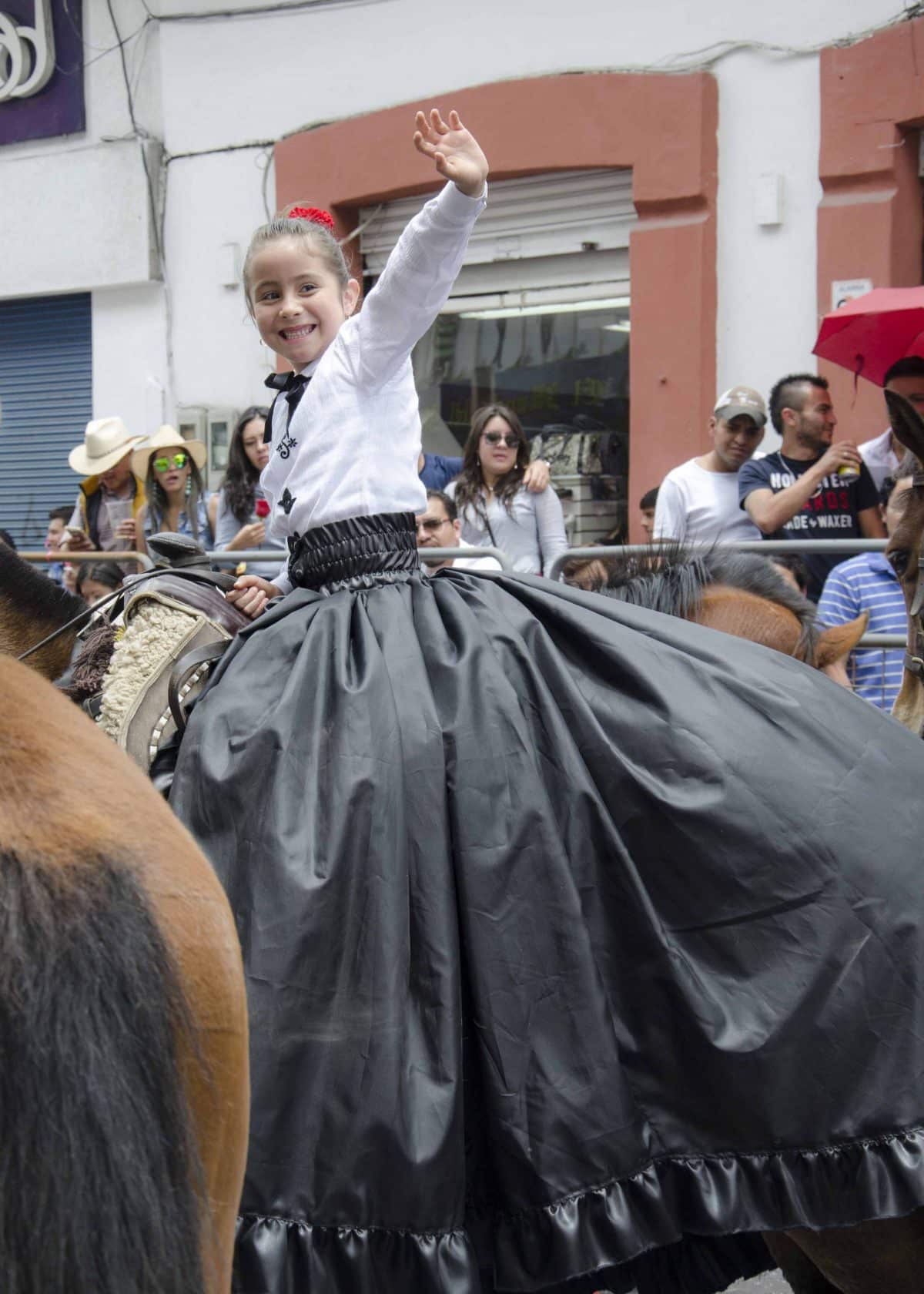 A young lady in full black skirt and white blouse turns to wave at someone she knows in the crowd