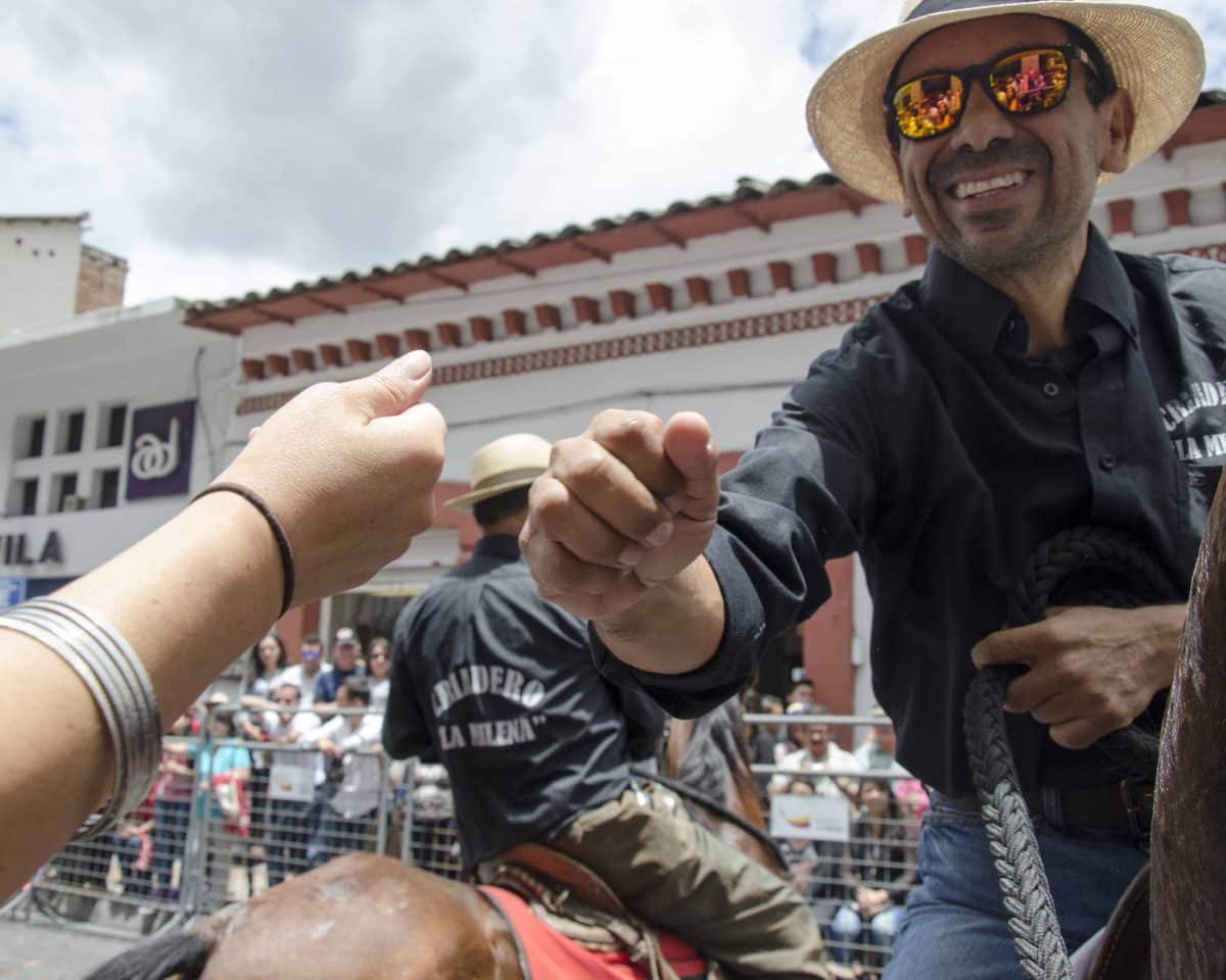 A man in sunglasses and straw hat hands a shot of a drink to a person watching the parade