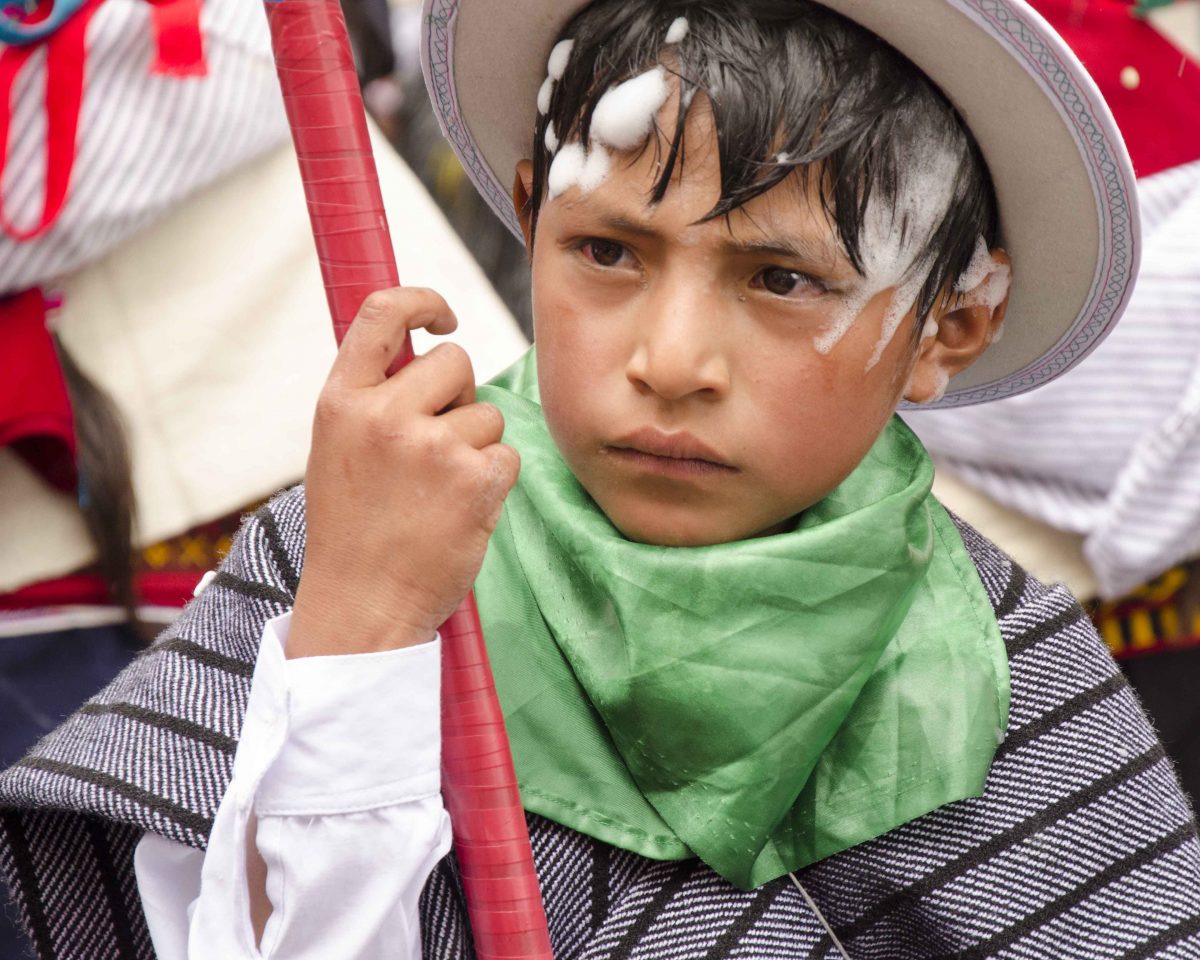 Un niño camina por la ruta del desfile; Carnaval en Guamote, Ecuador | ©Ángela Drake Joven con cara seria está vestido con poncho gris y sombrero de lana blanca