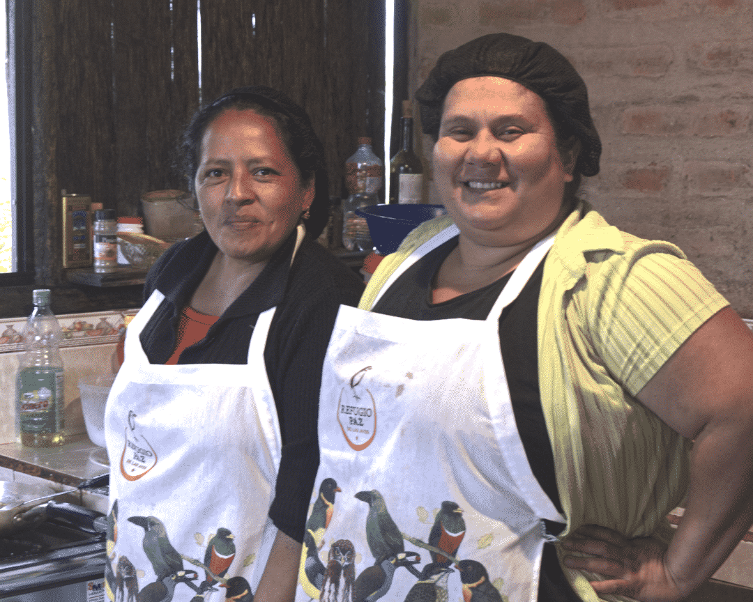 María Florcita y Diana Verá; refugio paz de las aves | ©Ángela Drake Dos mujeres sonrientes, vestidas con delantales con muchas imágenes de pájaros, están sonrientes en una cocina.
