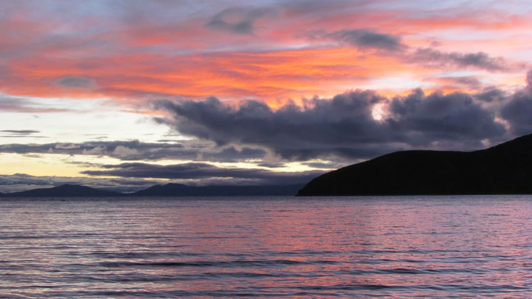 Un lago tranquilo refleja las nubes de color albaricoque, las nubes de tormenta más oscuras y el perfil negro profundo de una montaña.
