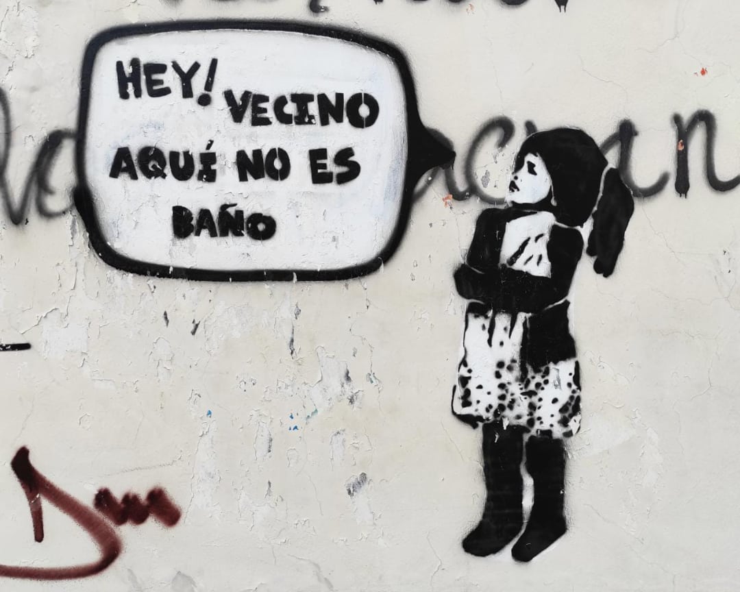 Un arte de sténcil, negro en blanco, una chica dice en estilo de cartoon, "HEY! VECINO AQUI NO ES BAÑO"