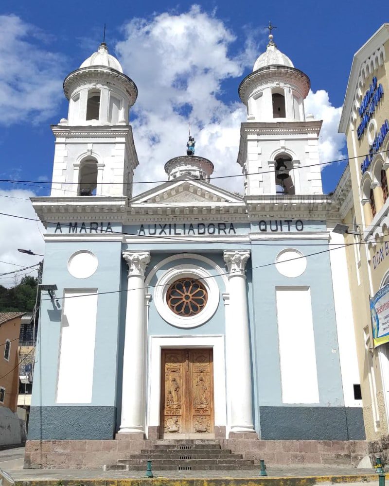 Una iglesia antigua tiene puerta de madera, un ventana rosada, dos torres blancos, y paredes pintadas de azul claro con acentos en blanco
