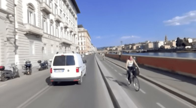 Una mujer en bicicleta se acerca al espectador por un carril bici a la derecha mientras seguimos a una furgoneta blanca por la carretera principal por delante