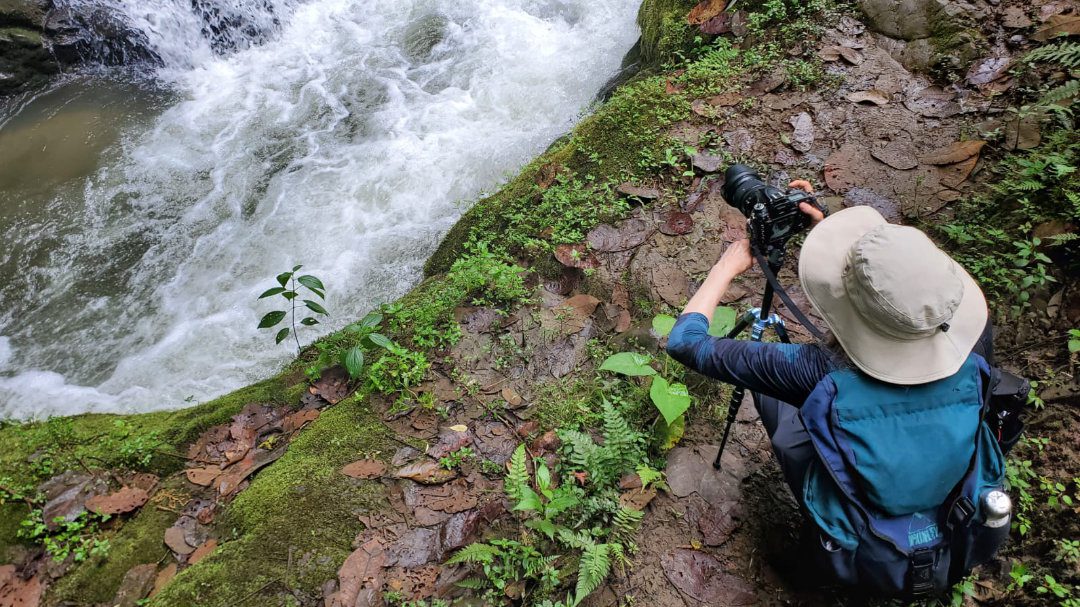 Angie con su equipo fotográfico, mochila y sombrero para el sol se las arregla para una exigente caminata en Ecuador | @Cliff Shakelford Una mujer se agacha sobre el trípode de una cámara mientras encuadra una foto de una cascada