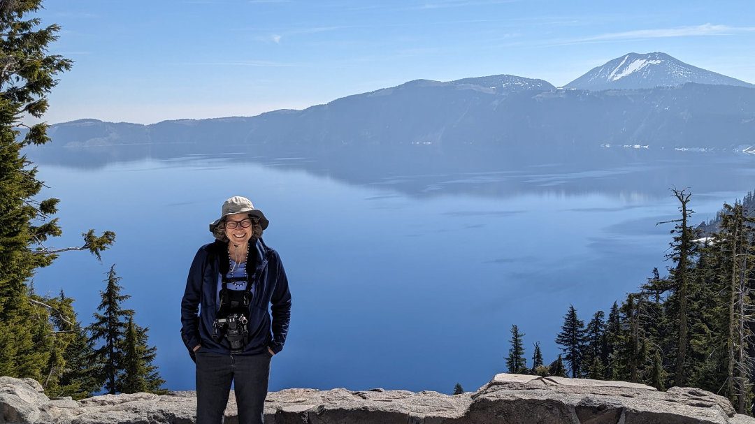 A women dressed in hiking gear and wearing a sunhat stands before the stunning blue Crater Lake in Oregon