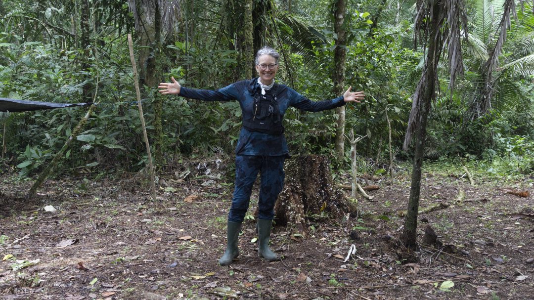 Angie está de pie con los brazos extendidos para mostrar su camisa de manga larga y pantalones largos metidos en botas de goma, medidas sugeridas para prevenir las picaduras de mosquito en la Amazonia