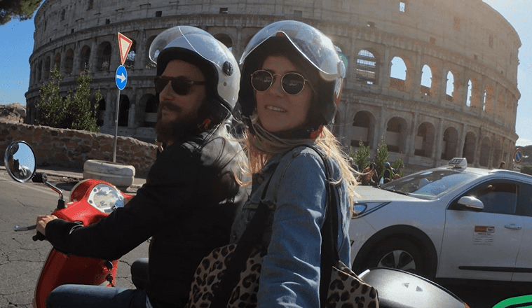 Un hombre y una mujer se toman un selfie sentados en un ciclomotor frente al Coliseo de Roma