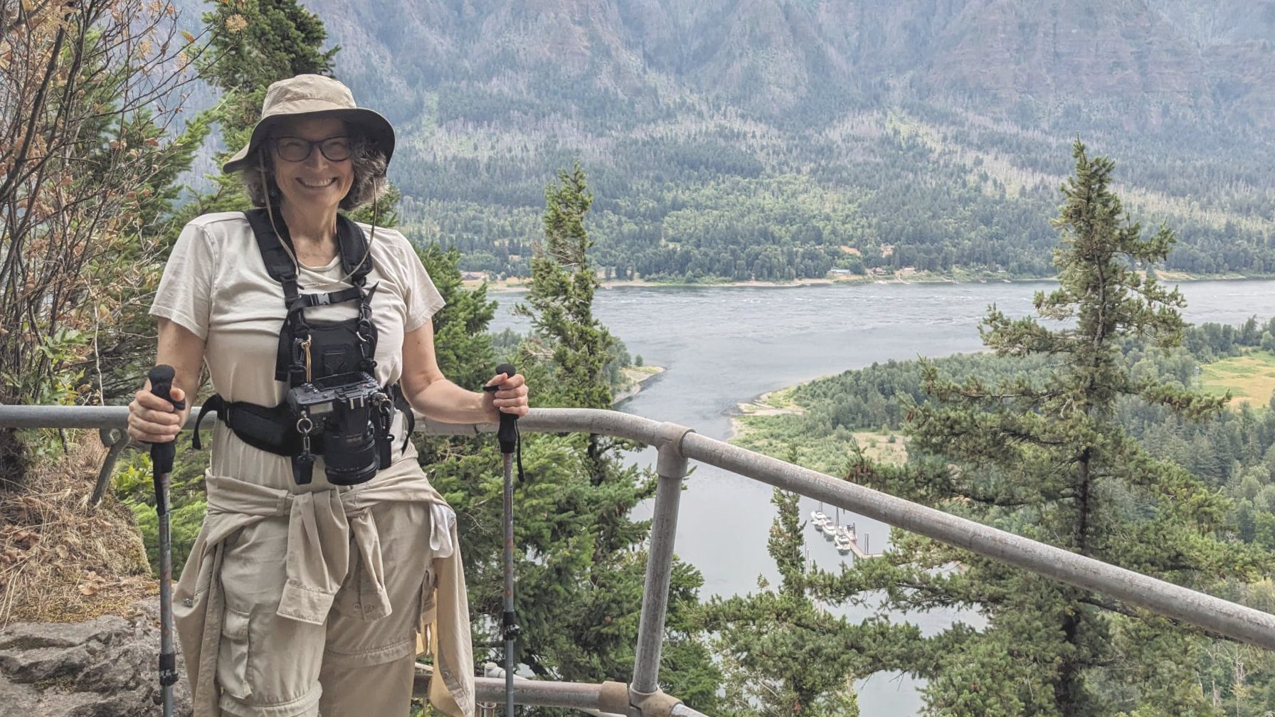 Angie utiliza bastones de senderismo en Beacon Rock, un sendero corto pero empinado a lo largo de la garganta del río Columbia, en Oregón | ©Ernest Scott Drake Una mujer vestida con equipo de senderismo y con un arnés de cámara sujeta unos bastones de senderismo mientras posa frente al río Columbia desde un sendero elevado.