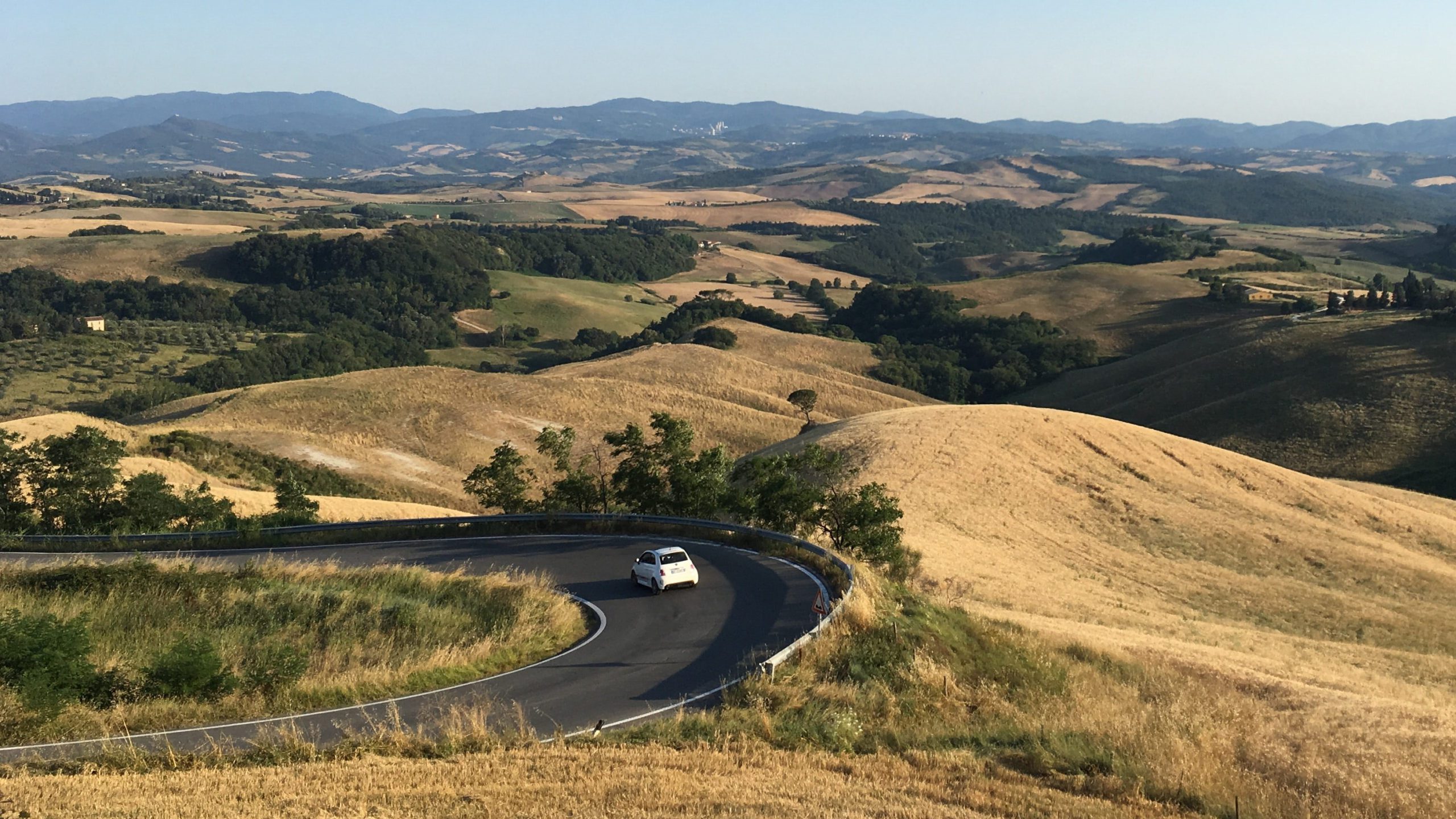 Un pequeño coche blanco que circula por Italia toma una curva cerrada en una carretera asfaltada con vistas a la impresionante campiña italiana.