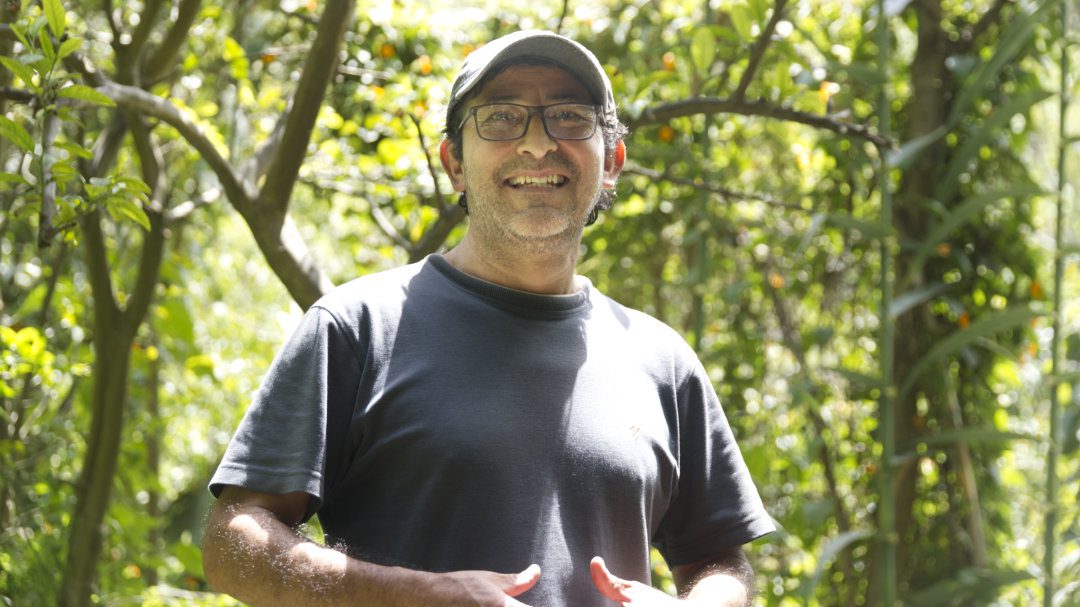 An Ecuadorian man in a dark tshirt and ball cap has a huge smile on his face as he stands in this garden at Pomasquinde