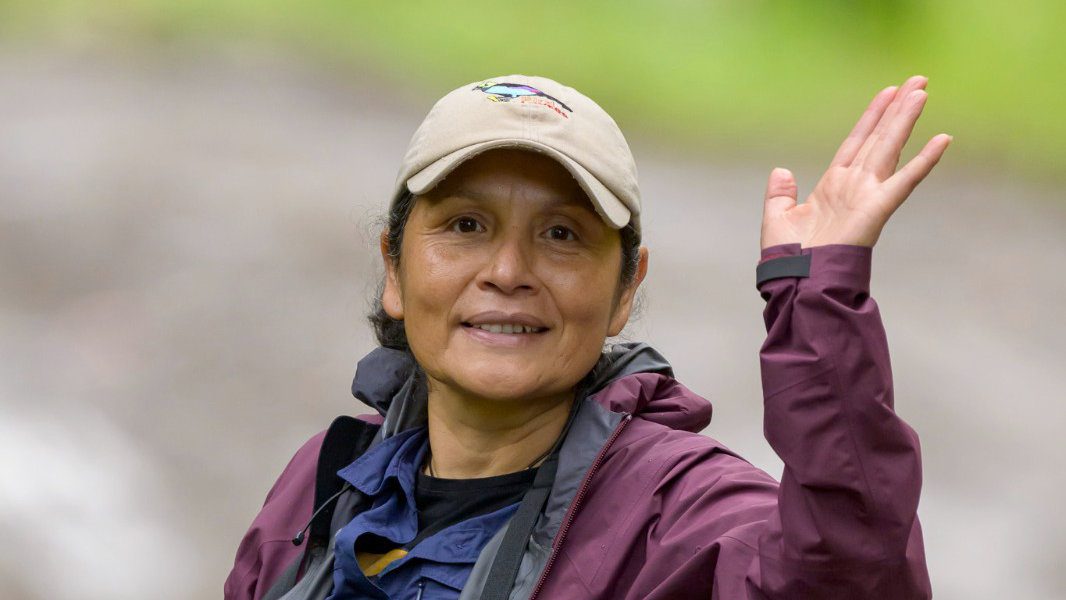 Close up of an woman wearing a ball cap and layered jackets waving at the camera