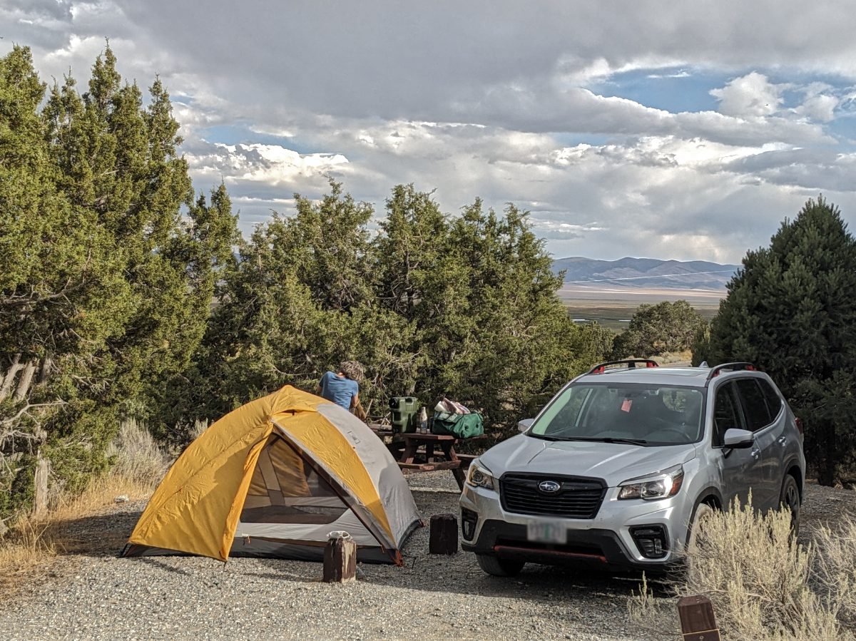 Automóvil de campamento en un piso de gravilla, vista de la carpa y el Subaru gris, mujer trabajando en la cocina del camping al fondo, todo ello con una magnífica vista del bosque de pinos, los humedales y las colinas lejanas.