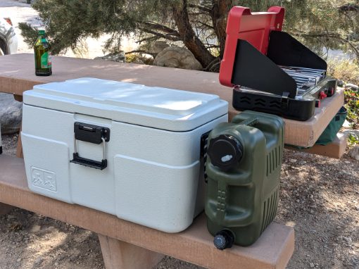 Our Rugged Roads Cooler sits on the bench of a picnic table alongside our water jug and near our campstove