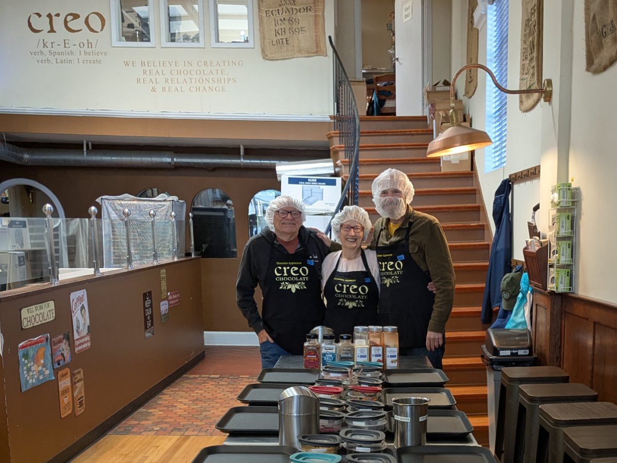 Tres personas están de pie al final de una larga mesa de preparación, vestidas con delantales de aprendiz de Creo, cofia para el pelo y una cofia para la barba de un caballero. 