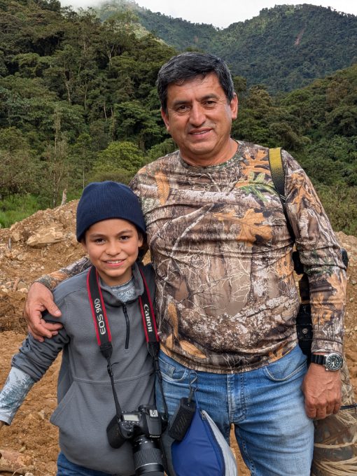 Un padre posa con el brazo alrededor de su hijo pequeño con las colinas y el bosque de fondo